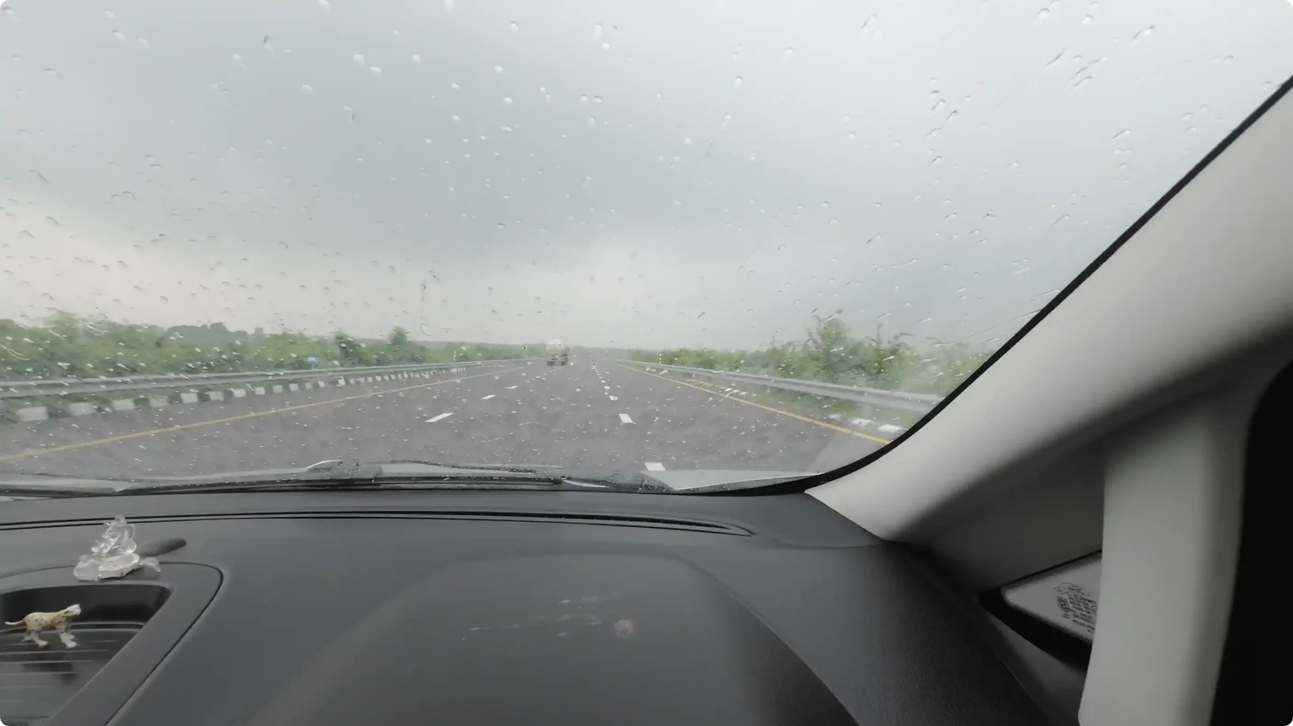 A dramatic driver's view from inside the car looking through a rain-streaked windshield at dark monsoon storm clouds on the Purvanchal Expressway