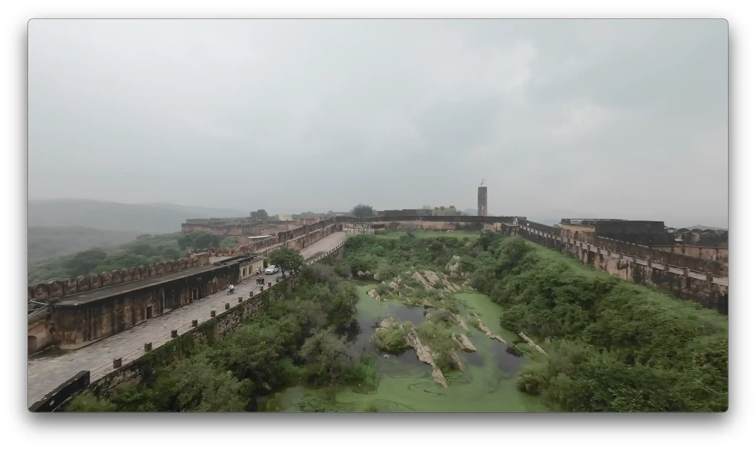 A wide view of the ancient stone walls, driving paths, and green overgrown water reservoirs inside Jaigarh Fort under a cloudy sky in Jaipur